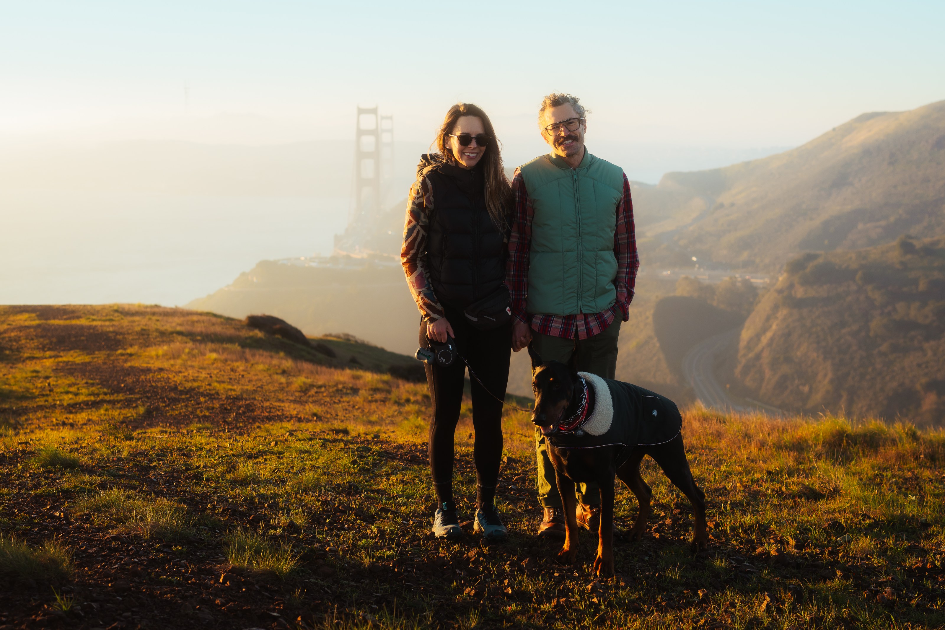 Anthony Palicci, his partner and their Doberman in the Marin Headlands, the Golden Gate Bridge in fog behind them